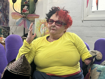 Kath Duncan has red hair and sits in her brightly decorated living room.